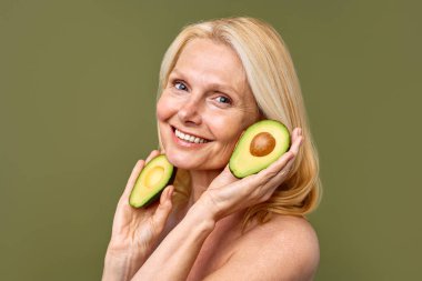 Olgun kadın, elinde avokado yarısıyla zeytin zemininde güven içinde poz veriyor. Kozmetoloji, cilt bakımı ve spa tedavileri. Kaldırma ve estetik ameliyat.