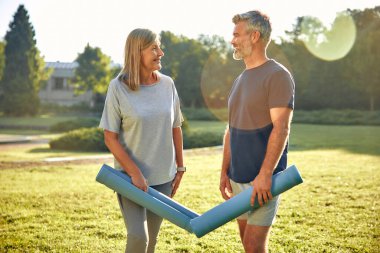 Neşeli ve mutlu bir çift, güzel güneşli bir parkta, yoga minderlerini heyecanlı bir şekilde tutuyor ve açık hava yoga seansına hazır, etraflarında canlı sağlık, enerji ve pozitiflik yayıyorlar.