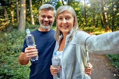 Neşeli bir çift canlı yeşil bir ormanın ortasında yürüyüş yaparken su şişelerini tutarak aktif yaşam tarzlarını ve çevrelerindeki doğal güzelliklerle derin bağlantılarını gösterirler.