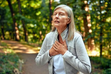 Görünüşte sıkıntılı bir yüzü olan bir kadın, doğanın güzelliği ile çevrili iken umutsuzca göğsünü sıkıyor, bir rahatsızlık, endişe ve duygusal mücadele anını canlandırıyor.