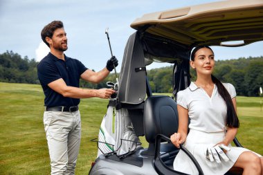 Manzaralı golf sahasında güneşli bir günde, bir erkek bir kadına golf arabasına yerleşmesi için yardım ediyor, boş zamanların ve açık hava aktivitelerinin özünü güzel bir şekilde yakalıyor.