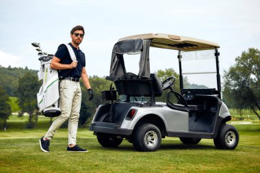 Golf sopalarını güvenli bir şekilde taşıyan bir adam canlı doğa ve renkli manzarayla çevrili iyi korunmuş bir golf sahasında arabasına doğru yürüyor ve huzurlu bir atmosfer yaratıyor.