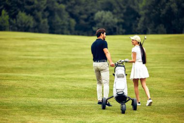 Şık bir çift, güzel manikürlü bir golf sahasında el ele dolaşır, golf sopalarını mutlu bir şekilde taşır ve sıcak güneş ışığında birlikte güzel vakit geçirmenin keyfini çıkarırlar.