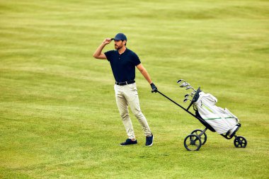 Rahat giyinmiş rahat bir golfçü, güzel bakımlı yemyeşil bir golf sahasında geziniyor, golf çantasına doğru itiyor ve golf oyununun tadını çıkarıyor.
