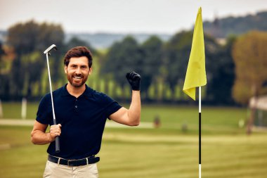 Siyah polo ve haki pantolonlu neşeli bir golfçü yeşil bayrağın yanında başarılı bir atış yaparak insanları bir araya getiren heyecanı kutluyor.
