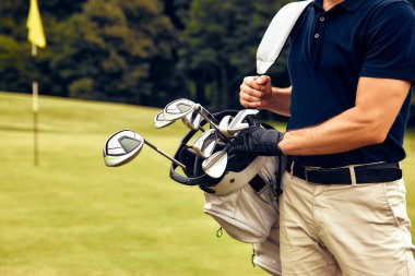 Yetenekli bir golfçü, elinde golf sopalarıyla yemyeşil alanda duruyor, heyecanlı bir güne hevesle hazırlanıyor, şık ve sportif bir görünüm sergiliyor.