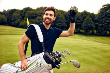 Heyecanlı, mutlu bir golfçü, golf sopalarını parlak ve güneşli bir golf sahasında tutarken sevinçle elini sallıyor. Sevdiği heyecanlı golf oyununa olan tutkusunu ve coşkusunu gururla sergiliyor.