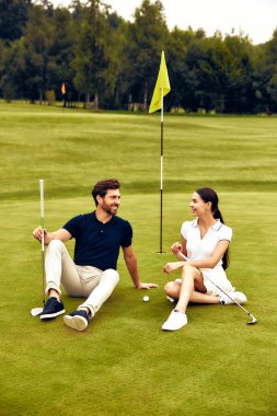 İki golfçü yeşilde oturmuş, neşeli bir konuşma yapıyor, açık havada eğleniyor ve birbirlerine eşlik ediyorlar.