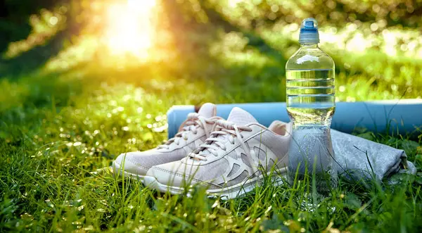 Bir çift şık ve şık koşu ayakkabısı, yeniden kullanılabilir bir su şişesi, ve düzgün sarılmış yoga minderi, hepsi canlı yemyeşil çimenlere yerleştirilmiş, doğada enerjik bir sabah egzersizi için ideal.