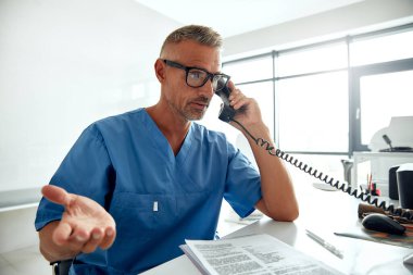 Aydınlık ve çağdaş bir ofis ortamında, ameliyat önlüğü giymiş odaklanmış bir doktor telefonla konuşuyor, aynı zamanda önemli tıbbi belge ve dosyaları da inceliyor.