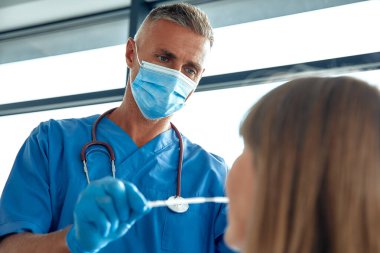 Tıbbi koruyucu eldivenli ve maskeli bir doktor klinikteki bir hastaya virüs testi yapıyor..