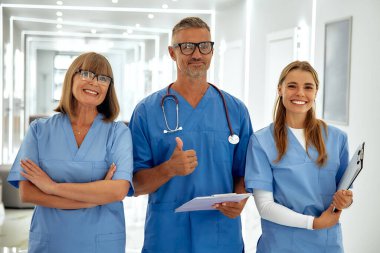 Parlak mavi önlüklü, gülümseyen üç kişiden oluşan özel bir sağlık ekibi, hastalara bir hastane koridorunda yardım etmeye hazır.