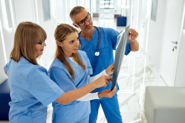 Yoğun bir hastane koridorunda, üç sağlık uzmanından oluşan bir ekip X-ray görüntülerini inceleyerek, ekip çalışmasının ve profesyonelliğin hasta sağlığındaki güvenliği açısından önemini vurguluyor.