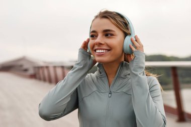 Şık kulaklıklı neşeli genç bir kadın dışarıda yürürken müzik ve çevresinden zevk alıyor, spor ve rahatlama zevkini yakalıyor, gevşemenin önemini vurguluyor.