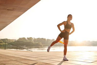 Kendini işine adamış ve tutkulu bir kadın güneş doğarken sakin nehir kenarındaki direnç bandını kullanarak canlandırıcı bir bacak egzersizi yapıyor. Fitness, sağlık ve genel canlılık ruhunu somutlaştırıyor.