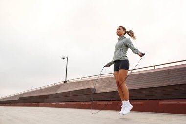 Formda bir genç kadın dışarıda egzersiz yapıyor, bir pistte ustalıkla ip atlıyor, sağlıklı bir yaşam tarzına ve genel sağlık ve zindelik için aktif spor faaliyetlerine olan bağlılığını gösteriyor.