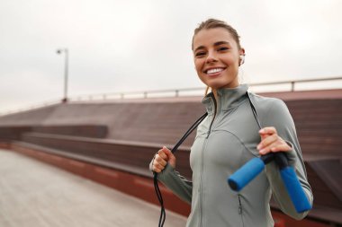 Gülümseyen genç bir kadın idman için tamamen hazır bir ip tutarken kendine güvenerek ayakta duruyor. Spor ve coşkunun özünü gerçekten somutlaştırıyor. Hepsi sakin bir arka planda duruyor.