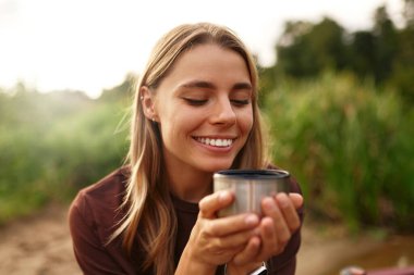 Canlı genç bir kadın neşe içinde huzurlu bir nehrin kenarında oturur, etrafındaki yemyeşil yemyeşil ortamda nazikçe bir yudum yudumlayarak, huzur, sükunet ve saf mutluluk hissi yayar.