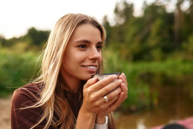 Canlı genç bir kadın neşe içinde huzurlu bir nehrin kenarında oturur, etrafındaki yemyeşil yemyeşil ortamda nazikçe bir yudum yudumlayarak, huzur, sükunet ve saf mutluluk hissi yayar.