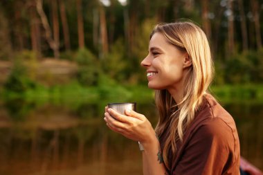 Genç bir kadın sakin bir gölün kenarında oturur, doğanın güzelliğiyle çevrili sıcak bir içkinin tadını çıkarırken, kendisi için huzurlu bir an geçirir, muhteşem manzaranın sunduğu huzurun tadını çıkarır.