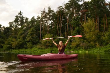 Neşeli bir kadın şen şakrak raketlerini kaldırırken sakin ve sakin bir gölde kano sürüyor. Etrafı tamamen yemyeşil ağaçlar ve doğa ile çevrili. Açık hava macerası ve doğa temaları için ideal.