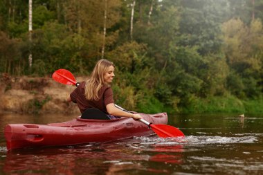 A young woman joyfully paddles her bright red kayak across the tranquil lake, embracing the stunning beauty of nature and creating unforgettable memories to cherish for a lifetime clipart