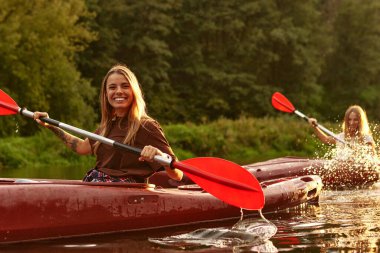 Büyüleyici bir günbatımı, iki neşeli kanocu, dingin ve huzurlu atmosferi geliştiren yemyeşil yapraklarla çevrili sakin sularda yüzerken güzel bir şekilde ortaya çıkıyor.