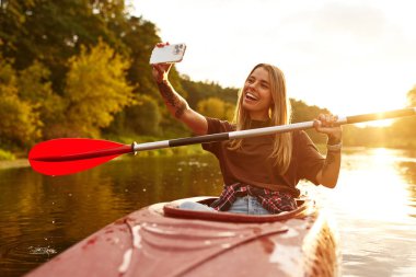 İnanılmaz mutlu bir birey, sakin bir nehirde kanoyla kayak yapıyor, büyüleyici bir günbatımı arkaplanı ile nefis bir selfie çekerken doğanın güzelliğinin ve maceranın heyecanının tadını çıkarıyor.