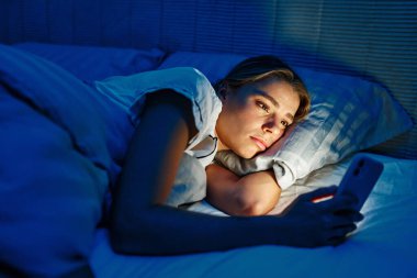 Gece çöktüğünde, genç bir kadın rahat yatağında teselliyi bulur, akıllı telefonuyla hafifçe aydınlatılır, günümüz dünyasını tanımlayan modern yaşamın ve dijital bağlantıların özünü yansıtır.
