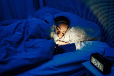Genç bir insan sıcak bir battaniyenin altında, akıllı telefonuyla huzurlu bir şekilde yatar. Ekrandaki yumuşak mavi ışık yüzlerini parlatacak, rahat bir gece sahnesi yaratacak.