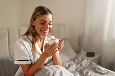 Memnun bir kadın, rahat yatağında sıcak bir fincan çayın keyfini çıkarır. Rahat bir sabah atmosferinde rahatlık ve rahatlık verir. Bu da onu huzur ve sükunetle sarar.