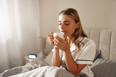 Memnun bir kadın, rahat yatağında sıcak bir fincan çayın keyfini çıkarır. Rahat bir sabah atmosferinde rahatlık ve rahatlık verir. Bu da onu huzur ve sükunetle sarar.