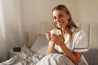 Memnun bir kadın, rahat yatağında sıcak bir fincan çayın keyfini çıkarır. Rahat bir sabah atmosferinde rahatlık ve rahatlık verir. Bu da onu huzur ve sükunetle sarar.