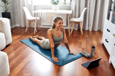 Genç bir kadın kendi evinde aktif olarak yoga yapıyor, sağlıklı rutininin hayati unsurları olarak sağlık ve sağlık durumunu vurgulayan online bir çalışma seansına katılıyor.