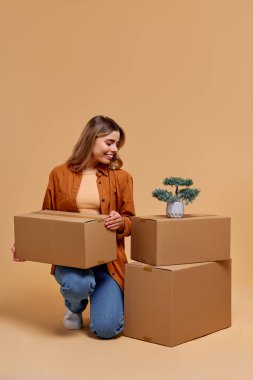 Neşeli bir atmosferde, neşeli genç bir kadın hareketliliği, ev dekorunu ve organizasyonu sembolize eden, sükuneti ve yaratıcılığı davet eden canlı bir saksı bitkisine hayran kalarak organize kutuların yanında diz çöker.