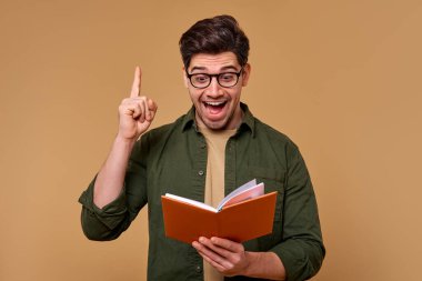 Gözlüklü neşeli bir genç adam neşeyle bir kitap tutuyor ve heyecanlı bir şekilde yukarı bakıyor. Bu resim eğitim, ilham ve motivasyon temalarıyla aynı hizaya gelir, bu da onu çeşitli bağlamlarda çok yönlü kılar.