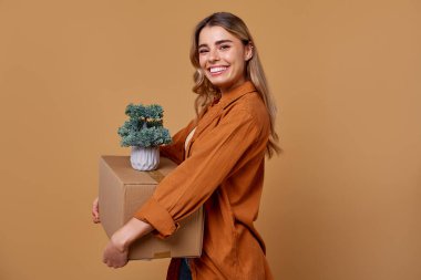 Neşeli genç bir kadın bir elinde canlı bir saksı bitkisi, diğer elinde sağlam bir karton kutu tutuyor. Bu da onun heyecan verici hareketini ve sonsuz olanaklarla dolu yeni bir başlangıcı simgeliyor.