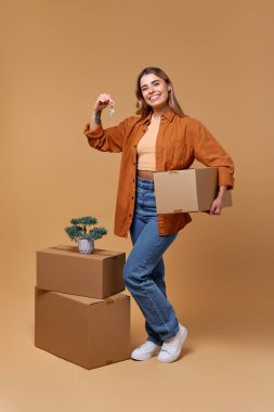 Kutu ve anahtarla neşeli genç bir kadın taşınan kutular ve canlı bir bitki ile çevrili, sıcak bir bej arka plan, somutlaştırıcı yaşam tarzı, yer değiştirme ve yeni başlangıçlar temaları karşı