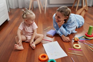 Neşeli bir anne, meraklı bebeğinin hayallerini canlandıran renkli oyuncaklarla çevrili sıcak ahşap bir zeminde canlı çizimler ve eğlenceli aktivitelerle yaratıcılığı keşfetmesini izler.