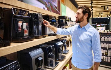 Bir adam, modern bir mağazada sergilenen çeşitli kahve makinelerini inceliyor ve inceliyor, yaklaşan alışverişi ve ihtiyaçlarına en uygun olan şeyleri inceliyor.