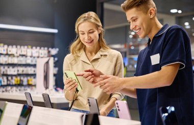 Müşteri, çeşitli teknolojilerle dolu canlı ve dinamik bir perakende ortamında çeşitli akıllı telefon seçeneklerini ayrıntılı bir şekilde incelerken bilgili bir satıcıya danışmanlık yapmaktadır.