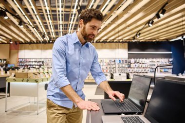 Moda bir elektronik butikte, genç bir adam çeşitli dizüstü bilgisayarları hevesle araştırıyor, modern teknolojiyi gözler önüne seriyor ve onların yenilikçi özellikleri ve işlevsellikleri için heyecanını dile getiriyor.