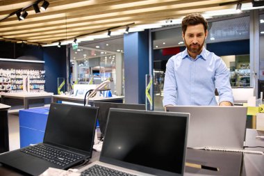 Moda bir elektronik butikte, genç bir adam çeşitli dizüstü bilgisayarları hevesle araştırıyor, modern teknolojiyi gözler önüne seriyor ve onların yenilikçi özellikleri ve işlevsellikleri için heyecanını dile getiriyor.
