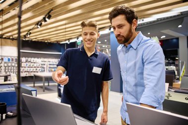 Bir elektronik mağazasının içinde, müşteri bir mağaza çalışanına ilgilendikleri dizüstü bilgisayarla ilgili bir konuşma yapmak için yaklaşır. Çalışan aygıt hakkında bilgi ve rehberlik sunar.
