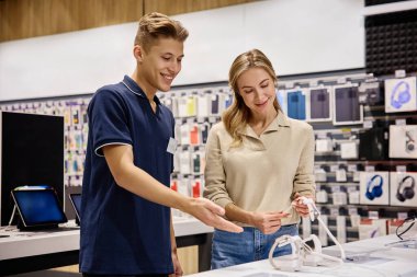 Bir müşteri, modern bir teknoloji mağazasında destekleyici ve arkadaş canlısı bir satıcıyla görüşür. En son cihazları, aksesuarları ve teknolojik yenilikleri zevkle araştırır.