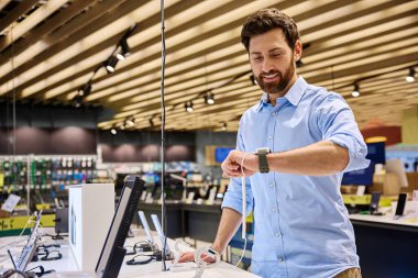 Bir adam canlı ve heyecan verici bir elektronik mağazasında sergilenen ve son teknoloji ve güncel perakende satış eğilimlerini sergileyen çeşitli akıllı saat yelpazelerini aktif olarak keşfediyor.
