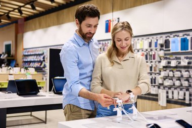 Genç bir çift, tabletler ve diğer moda cihazlar da dahil olmak üzere en son teknolojilerle dolu lüks, modern bir elektronik mağazasında çeşitli aletlerle neşeyle etkileşime geçerken görülüyor.