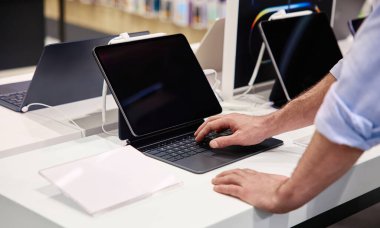 Müşteri titiz ve modern bir tableti inceliyor. Telaşlı bir elektronik mağazasında klavye ile donatılmış, çeşitli yüksek teknolojili cihazların satışa sunulduğu bir tablet.