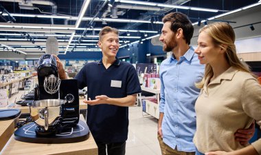 Meşgul bir perakende mağazasında, bilgili bir satış elemanı ilgili bir çifte yüksek teknoloji mutfak aletini göstererek, olumlu bir satın almayı teşvik etmek için eşsiz özelliklerini ve faydalarını vurgular.