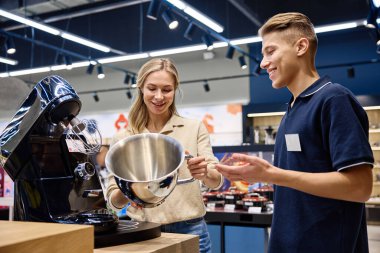 Meşgul bir perakende mağazasında, bilgili bir satış elemanı ilgili bir kadına yüksek teknoloji mutfak aletini göstererek, olumlu bir alışverişi teşvik etmek için benzersiz özelliklerini ve faydalarını vurgular.